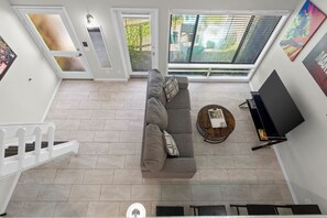 Un salon moderne avec un canapé gris, une table basse ronde en bois, un téléviseur à écran plat et une grande fenêtre équipée de stores.