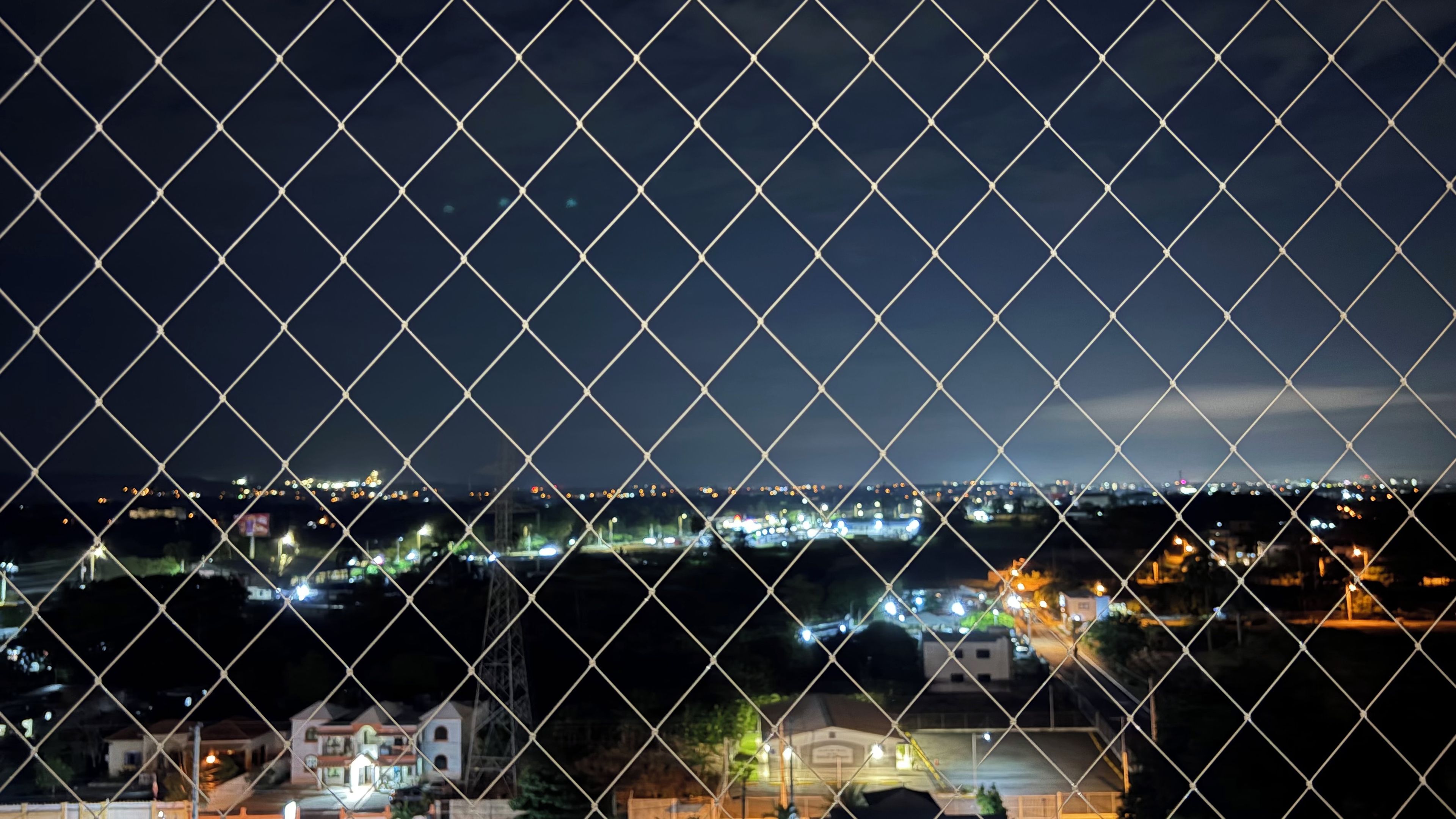 Une vue nocturne sur une ville, prise à travers une clôture en treillis métallique.