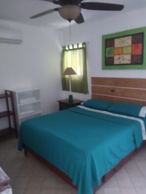 A bedroom with a bed, a ceiling fan, a nightstand, a lamp, a wall art piece, and a window with curtains. -  Encuentro Beach hidden gem / 1 Br, a/c, Internet, pool, backup electricity (Cabarete)