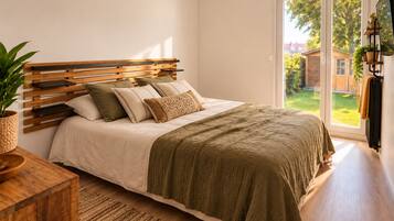 A bedroom with a wooden headboard, a bed with multiple pillows, a green throw blanket, a wooden bedside table with a plant, and a view of a garden through a sliding glass door.