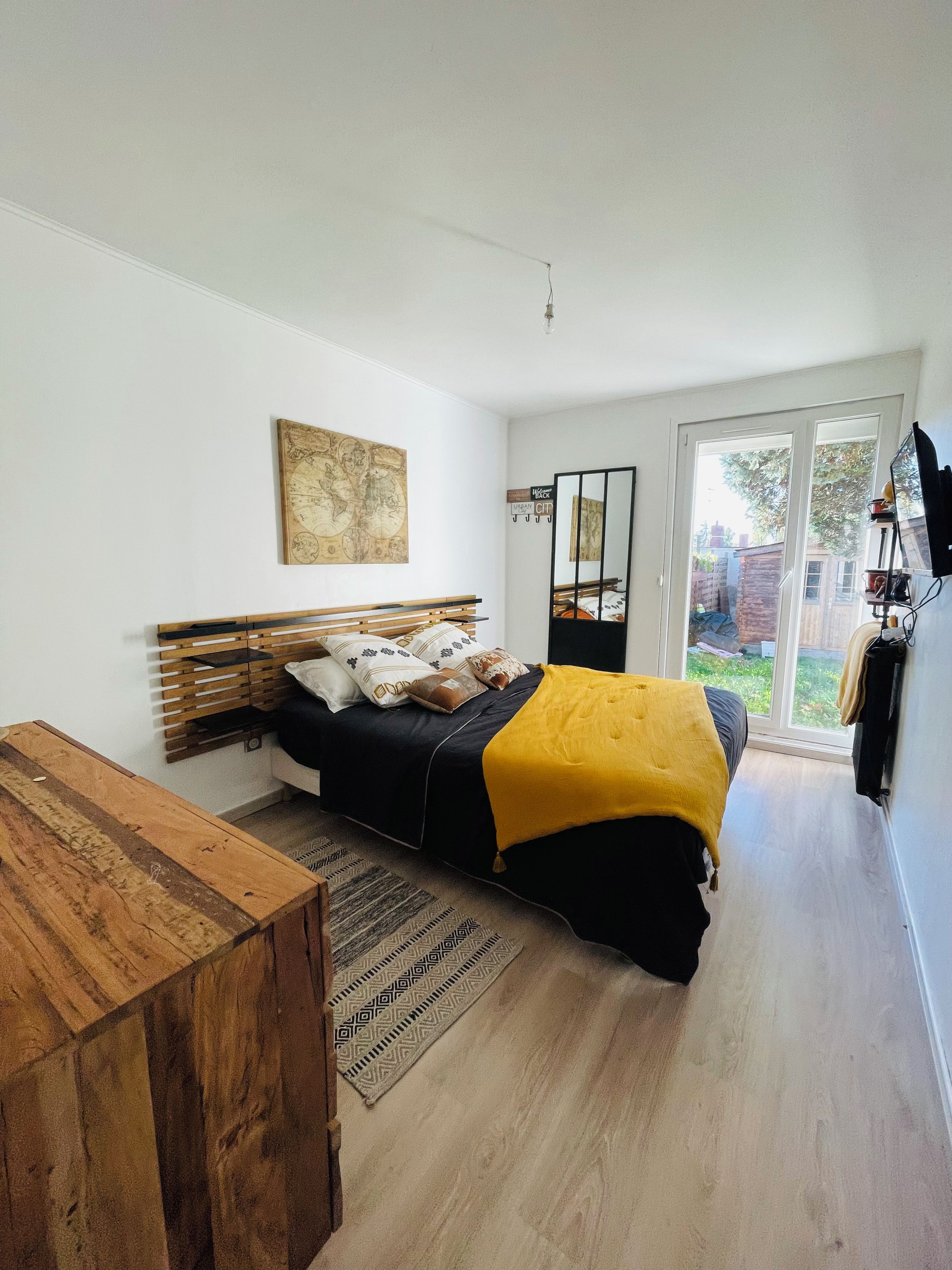 A bedroom with a bed, a wooden bedside table, a large window, and a view of the outdoors.