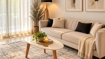 A modern living room with a beige sofa, a round coffee table with a plant and candles, a floor lamp, and abstract art on the wall.