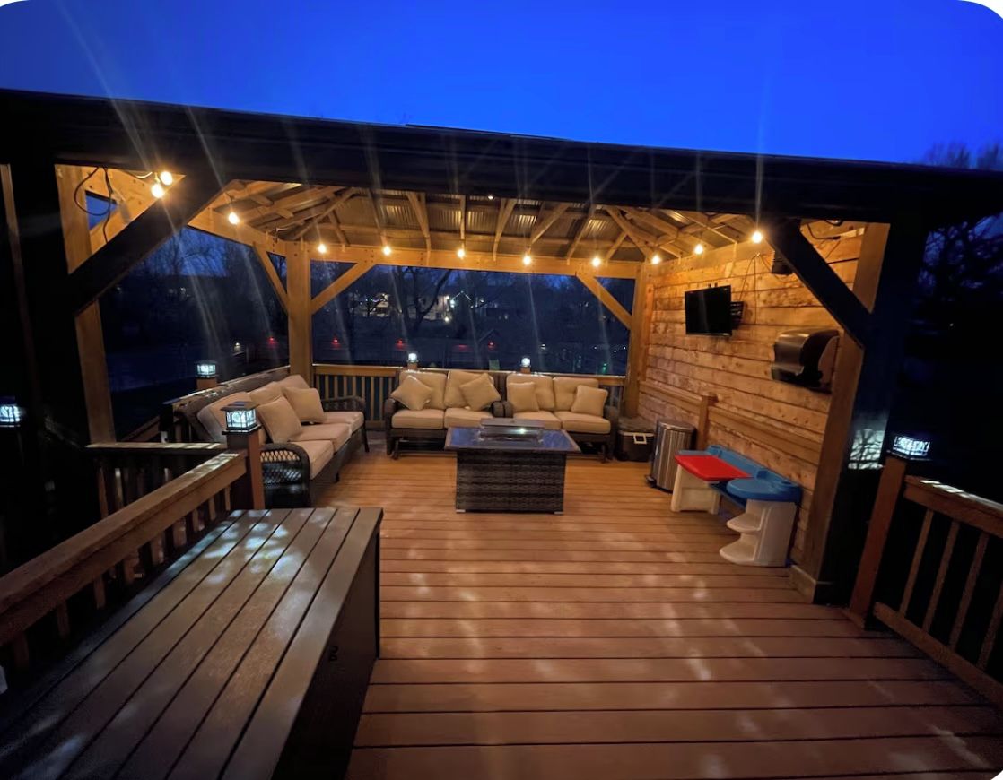 Une terrasse extérieure avec un plancher en bois, un espace salon couvert avec des canapés et une table basse, ainsi que des guirlandes lumineuses.