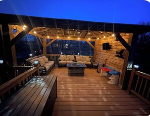 Une terrasse extérieure avec un plancher en bois, un espace salon couvert avec des canapés et une table basse, ainsi que des guirlandes lumineuses.