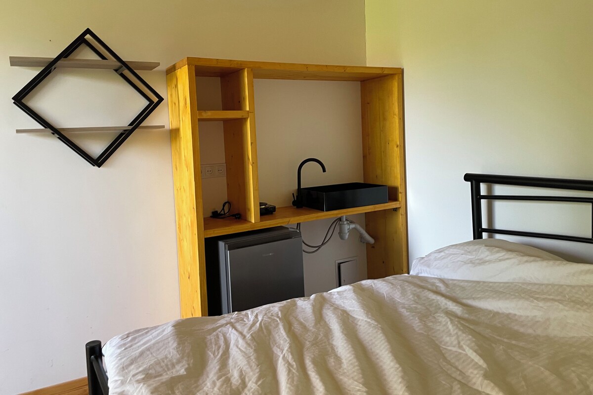 A bedroom with a bed, a wooden desk with a black sink, and a wall-mounted shelf.