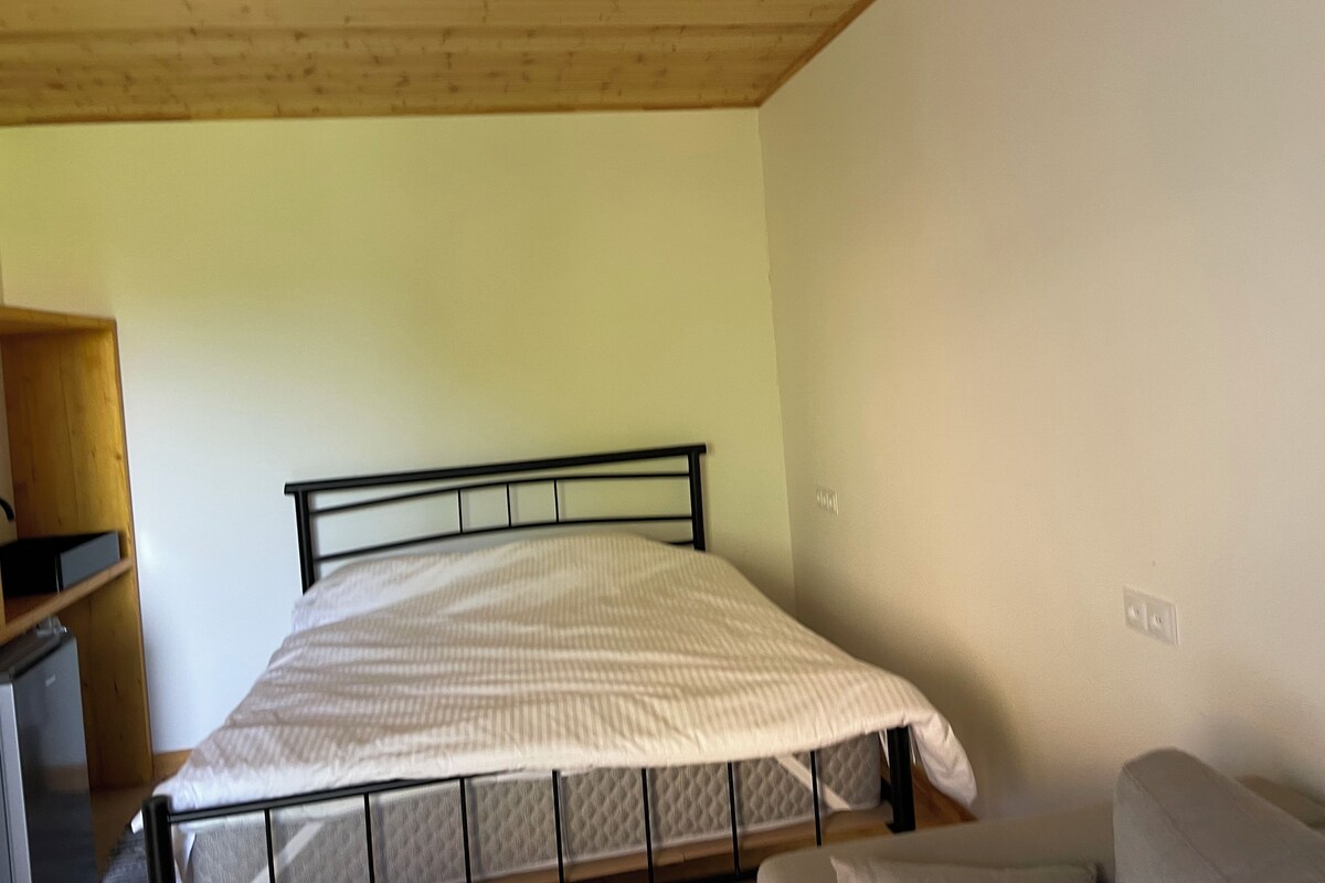 A single bed with a metal frame and a quilted mattress, a wooden shelf unit, and a light-colored wall.