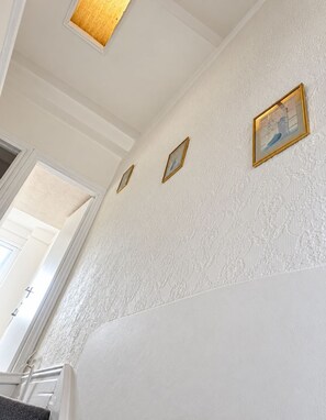 Un couloir aux murs blancs ornés d’un motif texturé, avec un plafond en bois et trois tableaux encadrés accrochés au mur.