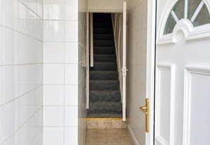 A hallway with white tiled walls, a dark carpeted staircase, and a door with a brass handle. - 1 Bed Flat Sleeps 2 - Pets Welcome - Tv & WiFi (Middlesbrough)