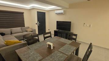 A living room with a sofa, dining table, chairs, a TV, and an air conditioning unit.