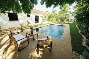 Una piscina con borde de azulejos, rodeada por un jardín con diversas plantas y césped. Hay dos sillas de mimbre y una mesa con cojines.