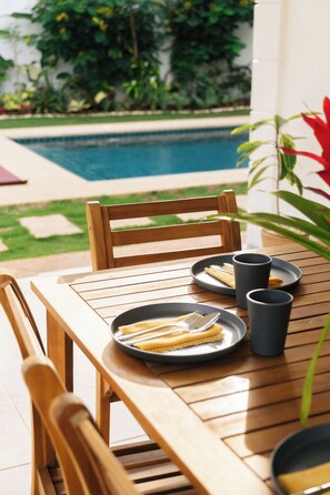 Una mesa de madera con platos, cubiertos y vasos, colocada al aire libre con una piscina y follaje al fondo.