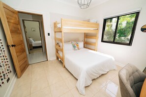 Una habitación con literas, una silla, un ventanal con vistas a zonas verdes y una puerta que da a otra habitación.