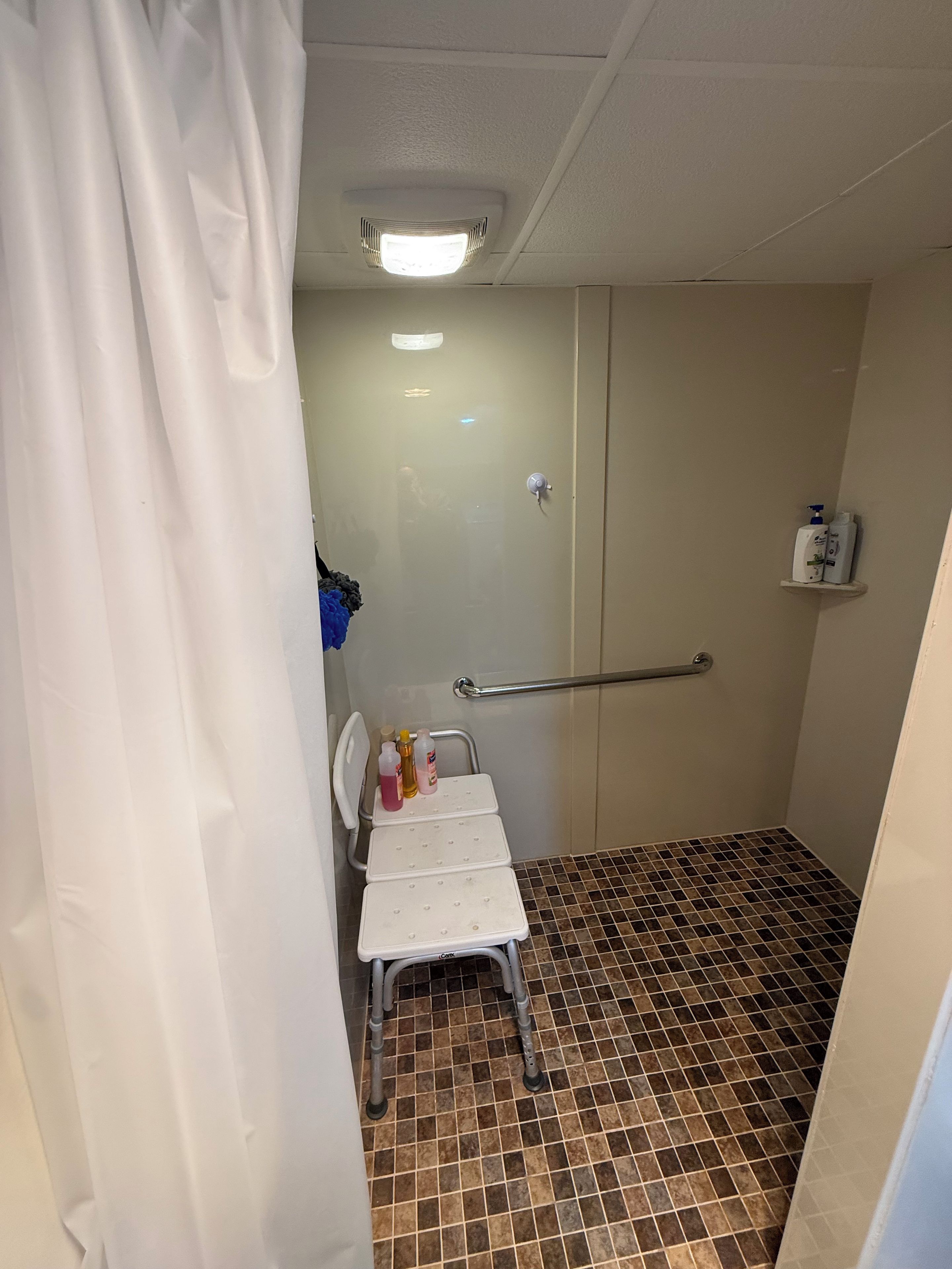 Een kleine badkamer met een douche, een witte stoel, een handgreep en een plank met toiletartikelen.
