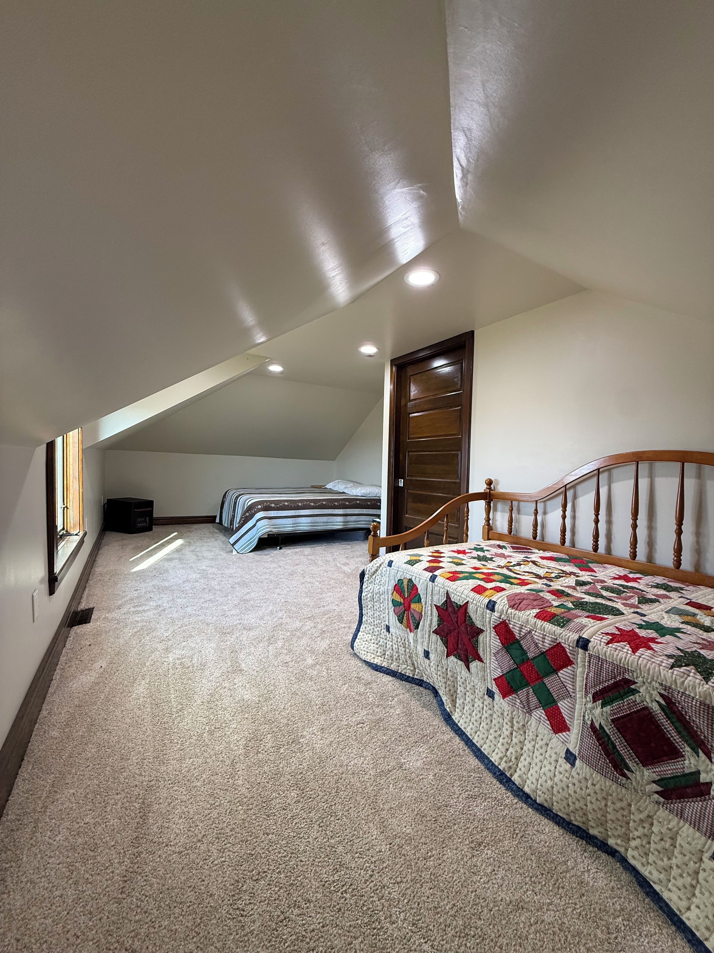 A bedroom with a sloped ceiling, a wooden bed frame, a quilt, and a window.