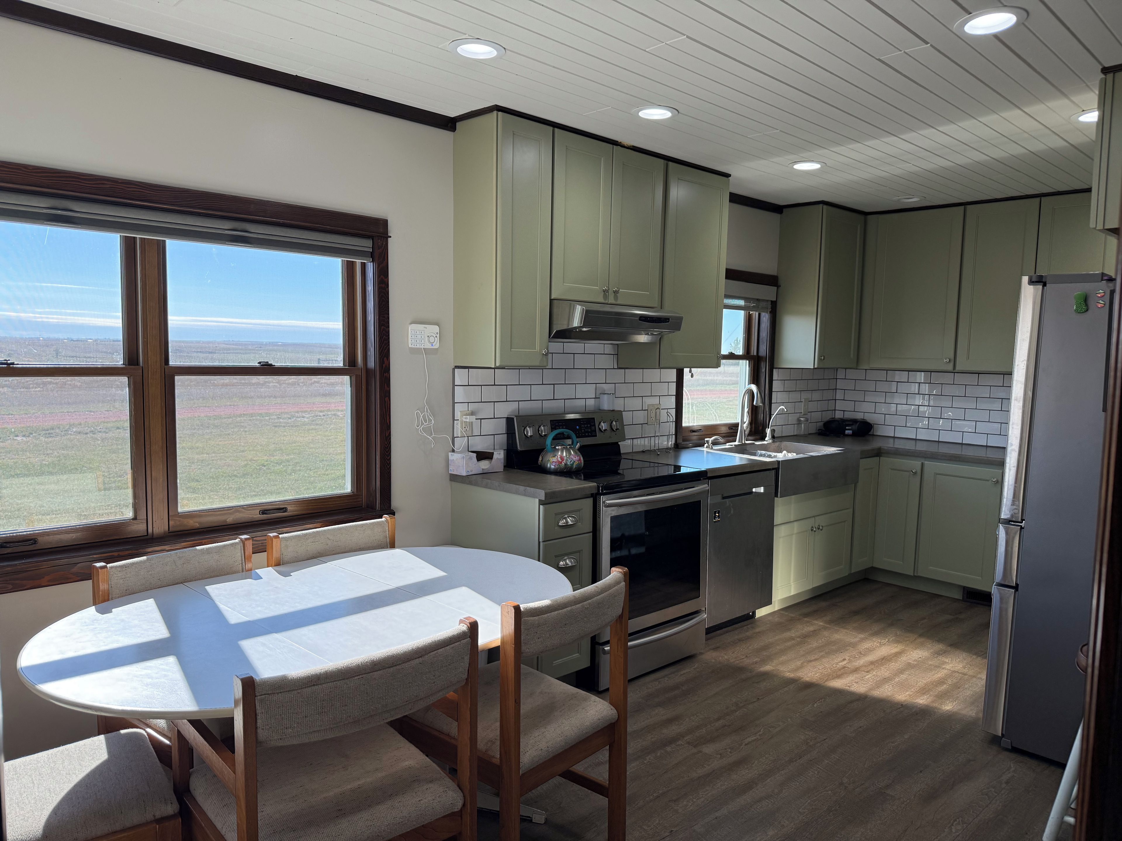 A kitchen with a dining table and chairs, a view through the window, and a refrigerator.