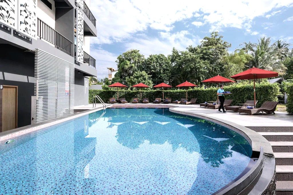 A swimming pool with lounge chairs and umbrellas, surrounded by greenery and a building with balconies.