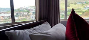 Uma cama com roupa de cama branca e um travesseiro vermelho, uma janela com cortinas e vista para uma área residencial.