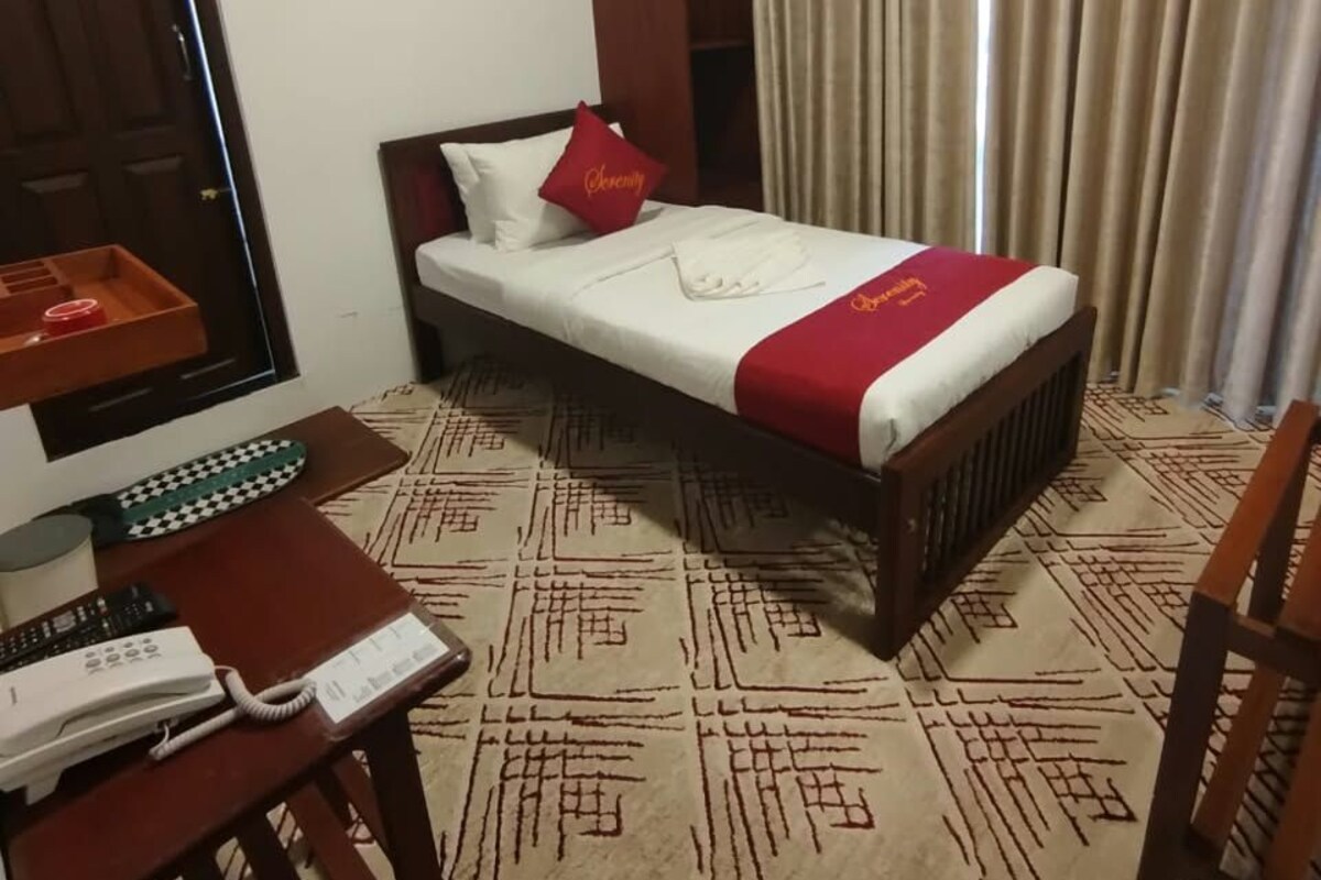Quarto de hotel com cama, escrivaninha com telefone, cadeira, quadro na parede e janela com cortinas.