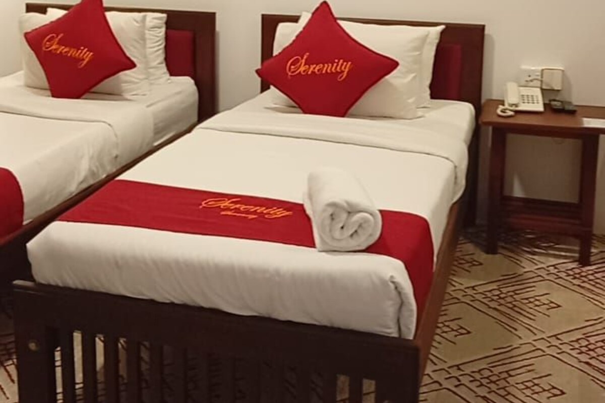 Quarto de hotel com duas camas, cada uma com um travesseiro vermelho e um edredom branco. Há uma pintura na parede acima das camas e um telefone na mesa de cabeceira.
