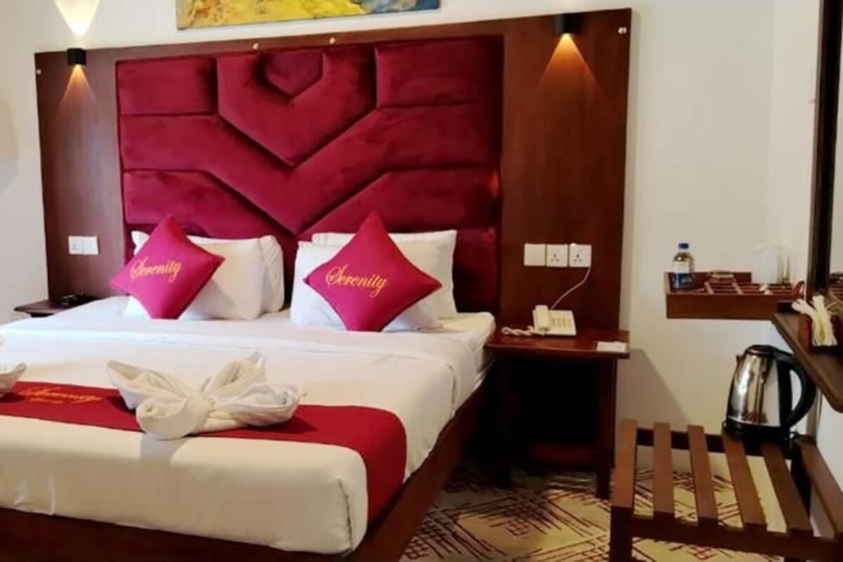 Habitació d'hotel amb un llit gran, dos coixins amb la paraula 'Serenity', una taula de nit amb una làmpada i una petita estant amb objectes.