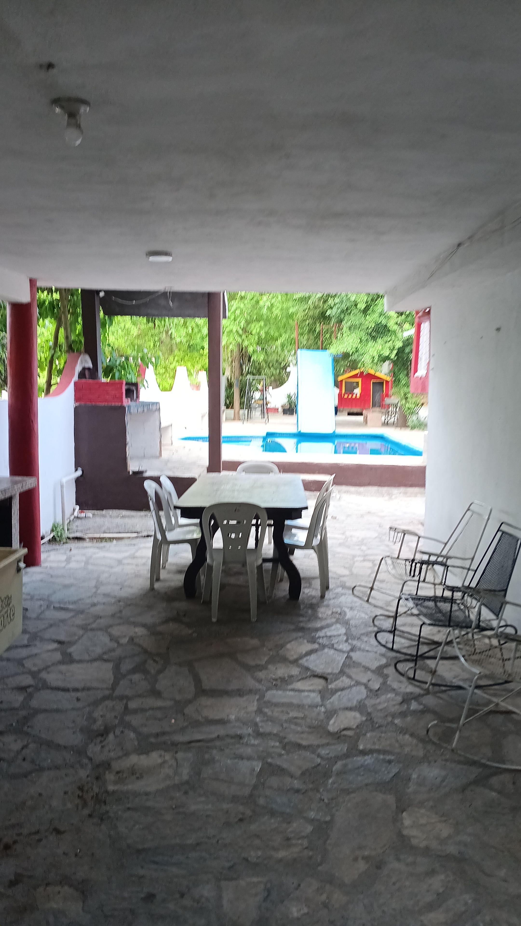 An outdoor patio area with a stone floor, a table with chairs, and a pool in the background.