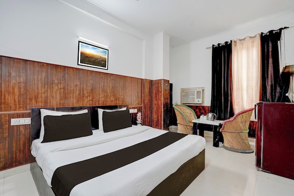 Habitación de hotel con cabecera de madera, cama con ropa de cama blanca y oscura, una mesita con una tetera, una silla de mimbre y un armario rojo.