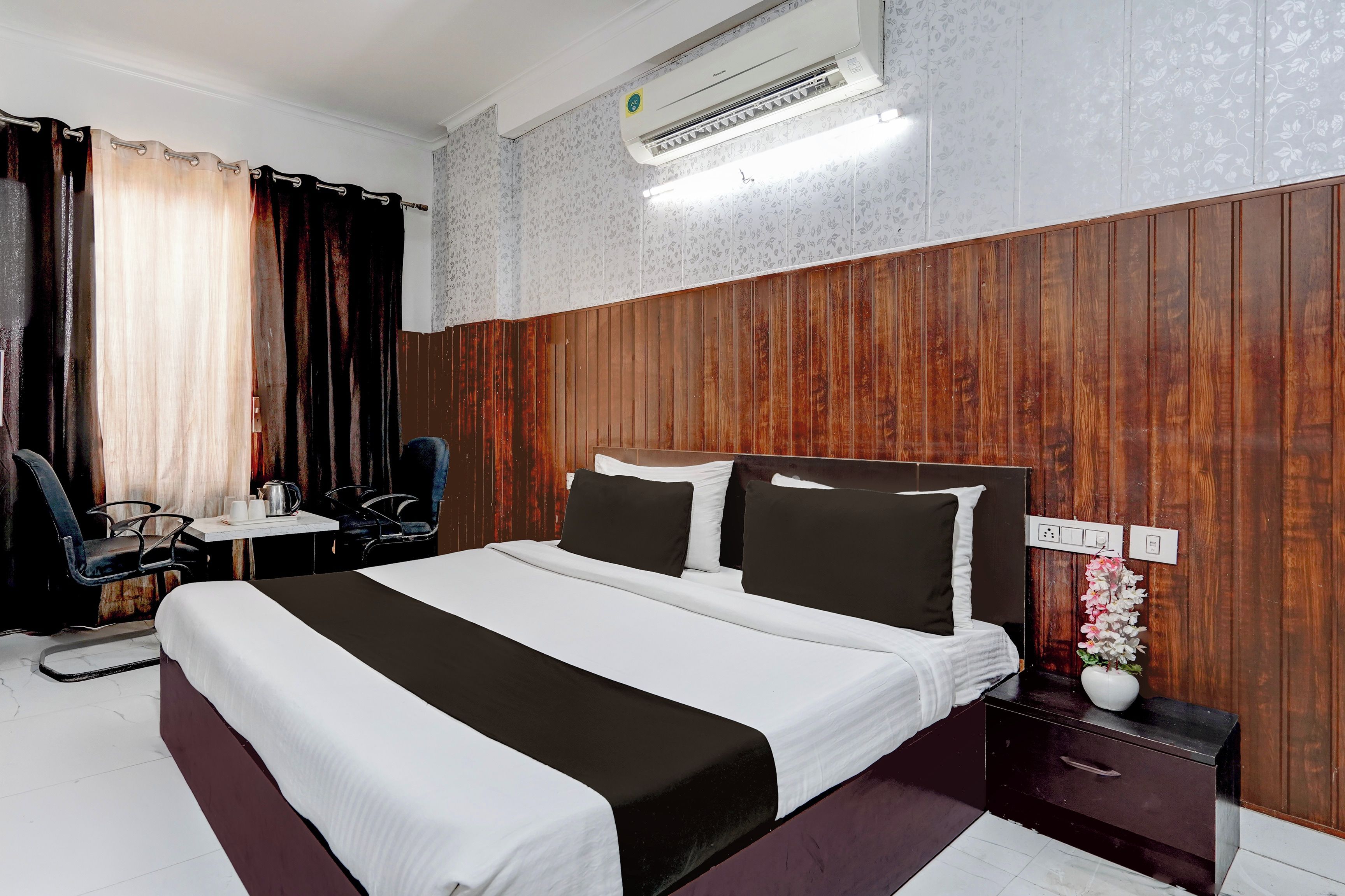 A hotel room with a bed, a nightstand, a desk with chairs, and an air conditioning unit.