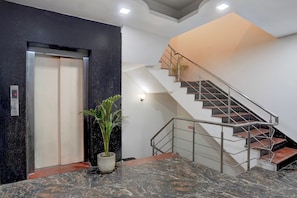 Un interior con piso de mármol, una planta en maceta, una escalera con barandillas de metal y un ascensor.