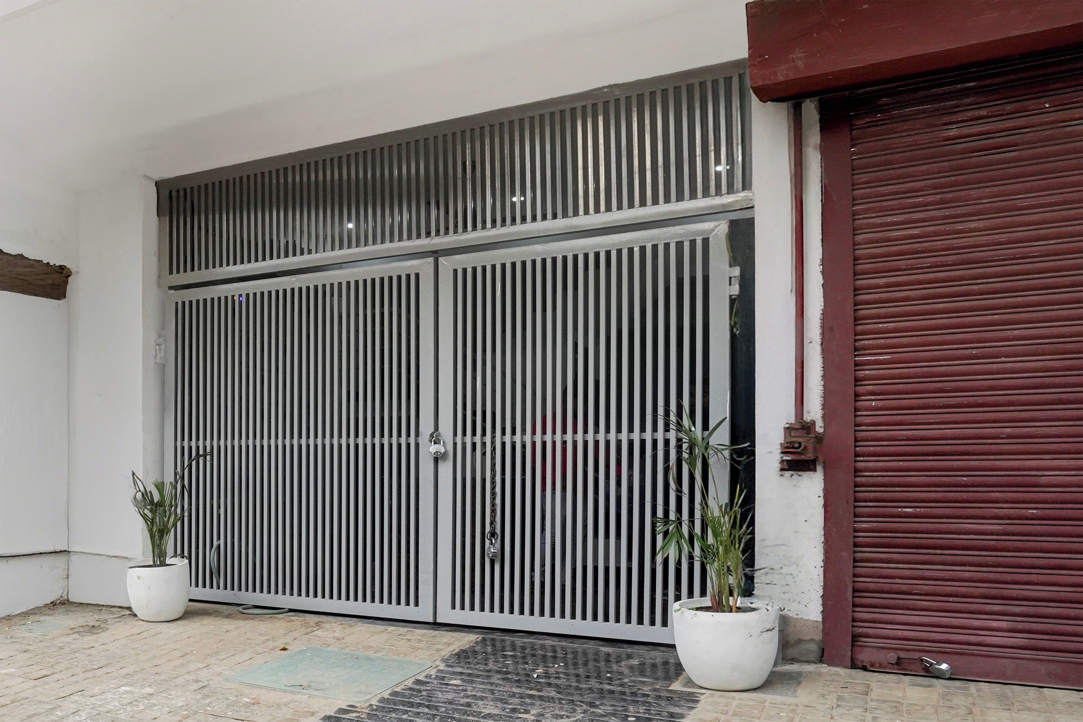 Una reja metálica cerrada con diseño de barras verticales, una puerta de garaje roja y dos macetas con plantas en el suelo.