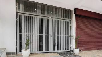 Una entrada protegida por un portón metálico, con dos plantas en macetas y un acceso con contraventanas rojas.