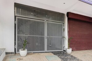 Una entrada protegida por un portón metálico, con dos plantas en macetas y un acceso con contraventanas rojas.