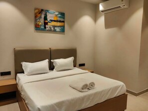 A hotel room with a bed, two pillows, a folded towel, a nightstand, an air conditioning unit, and a painting on the wall. - Hotel O R K Residency (Gurgaon)