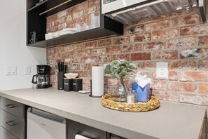 A kitchen with a brick wall, a coffee maker, a sugar container, a napkin holder, and a basket with a plant and tissues. - SoHo Vibes - EaDo Lofts #B (Houston)
