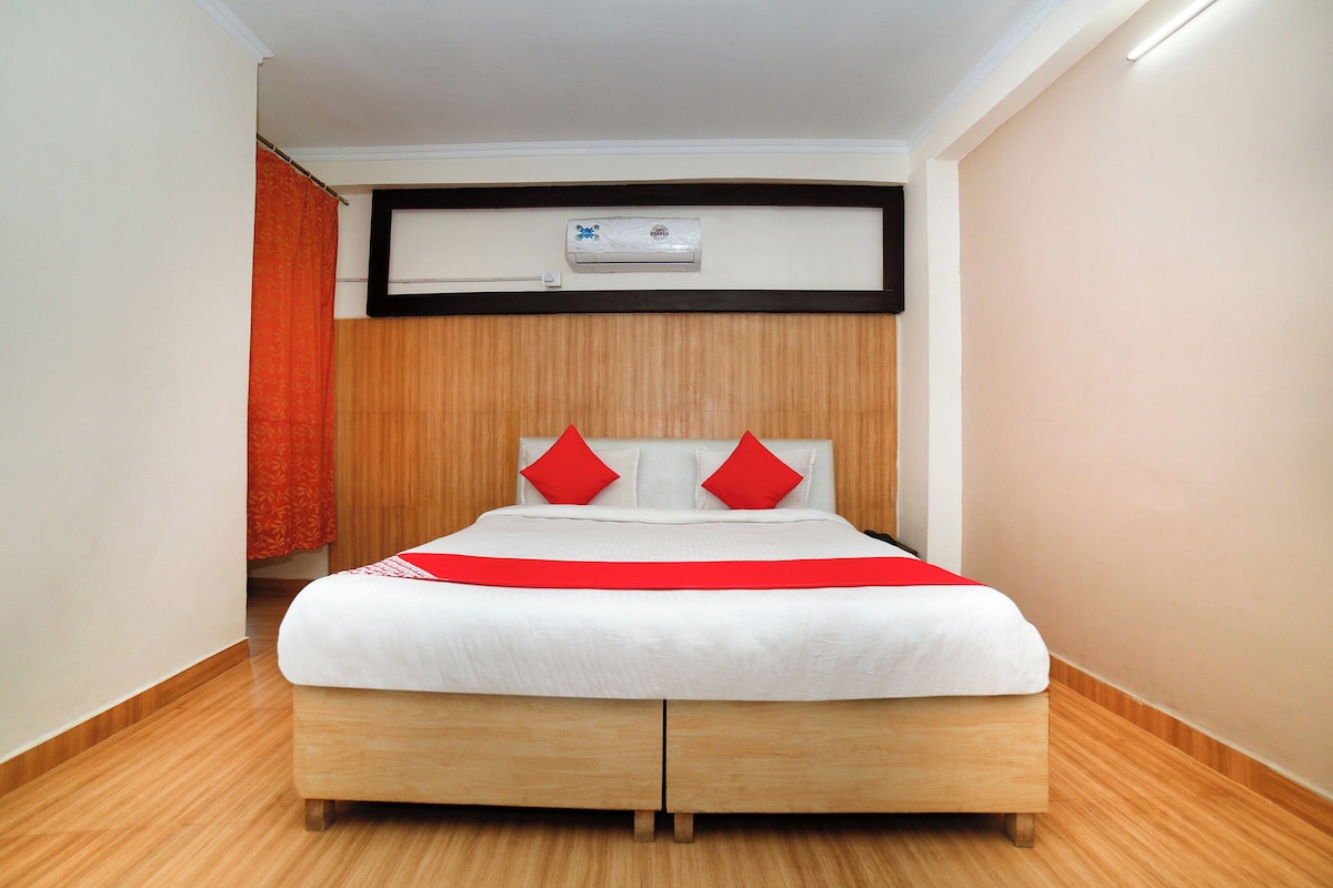 A hotel room with a bed, wooden flooring, a white wall, and a ceiling-mounted air conditioning unit.