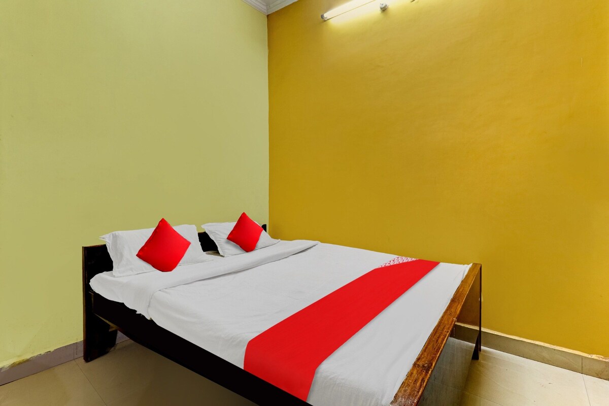 Una cama individual con ropa de cama blanca y detalles rojos, ubicada frente a una pared amarilla con una lámpara colgante.