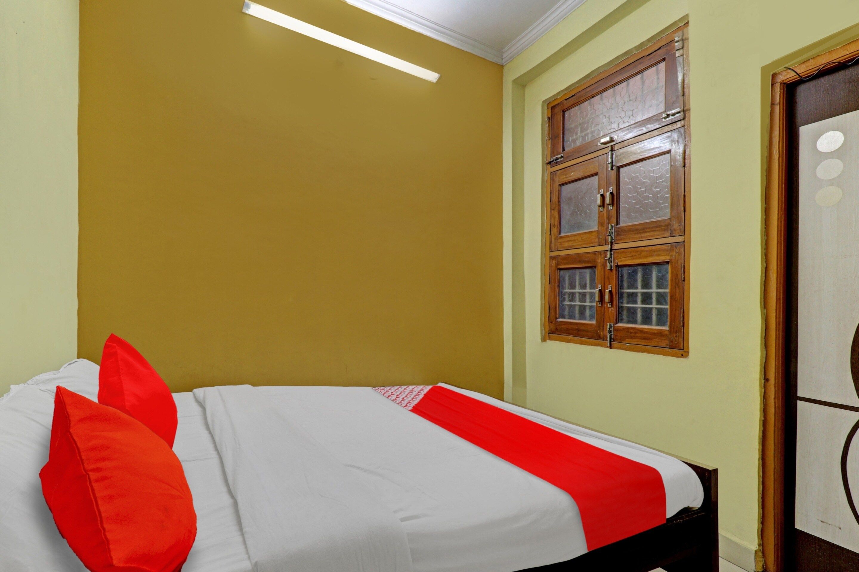 Una cama individual con colcha blanca y roja, cabecera de madera y una ventana con marco y vidrios de madera.