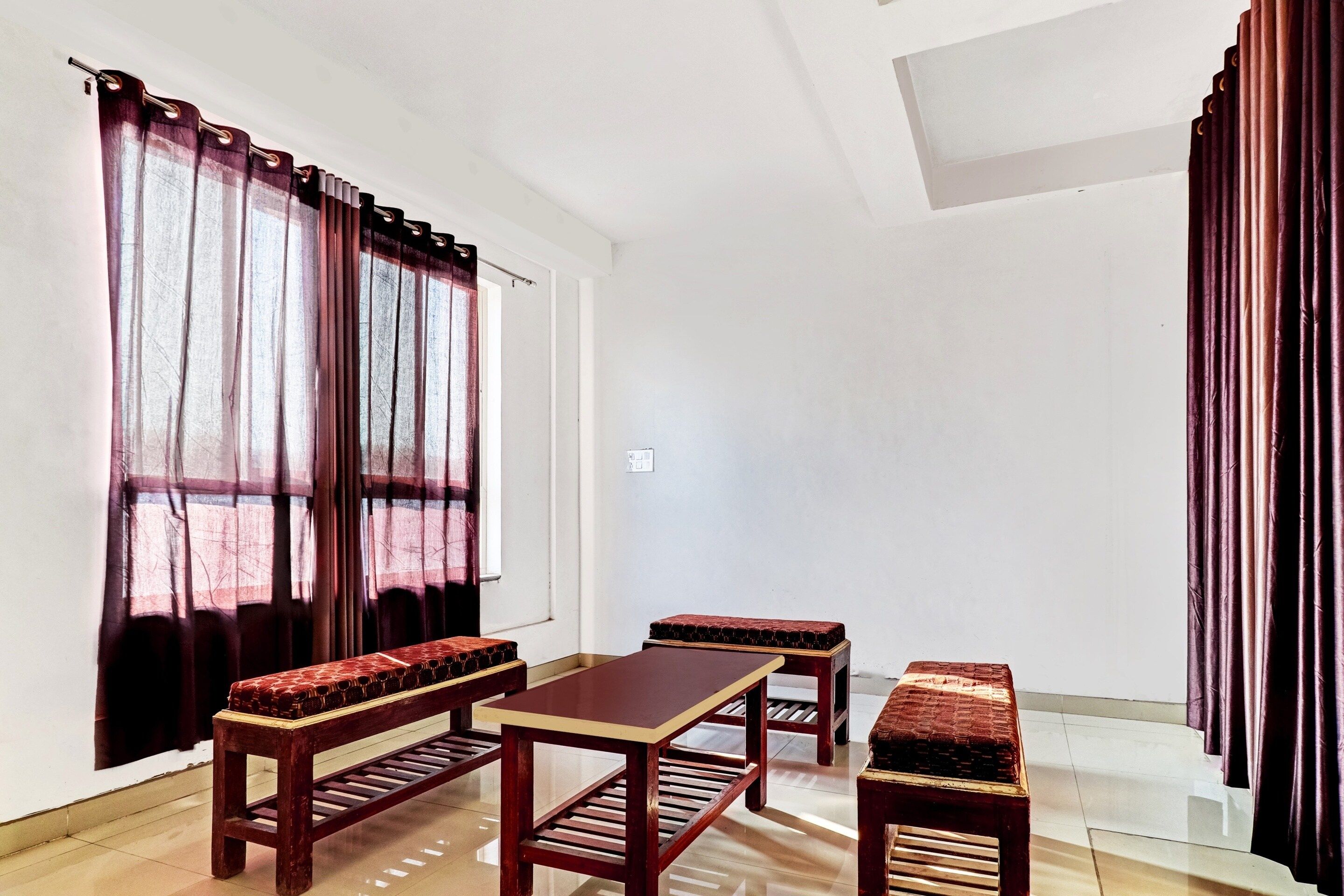 Una habitación con una mesa de madera y bancos, ventanas amplias con cortinas y suelo de baldosas.