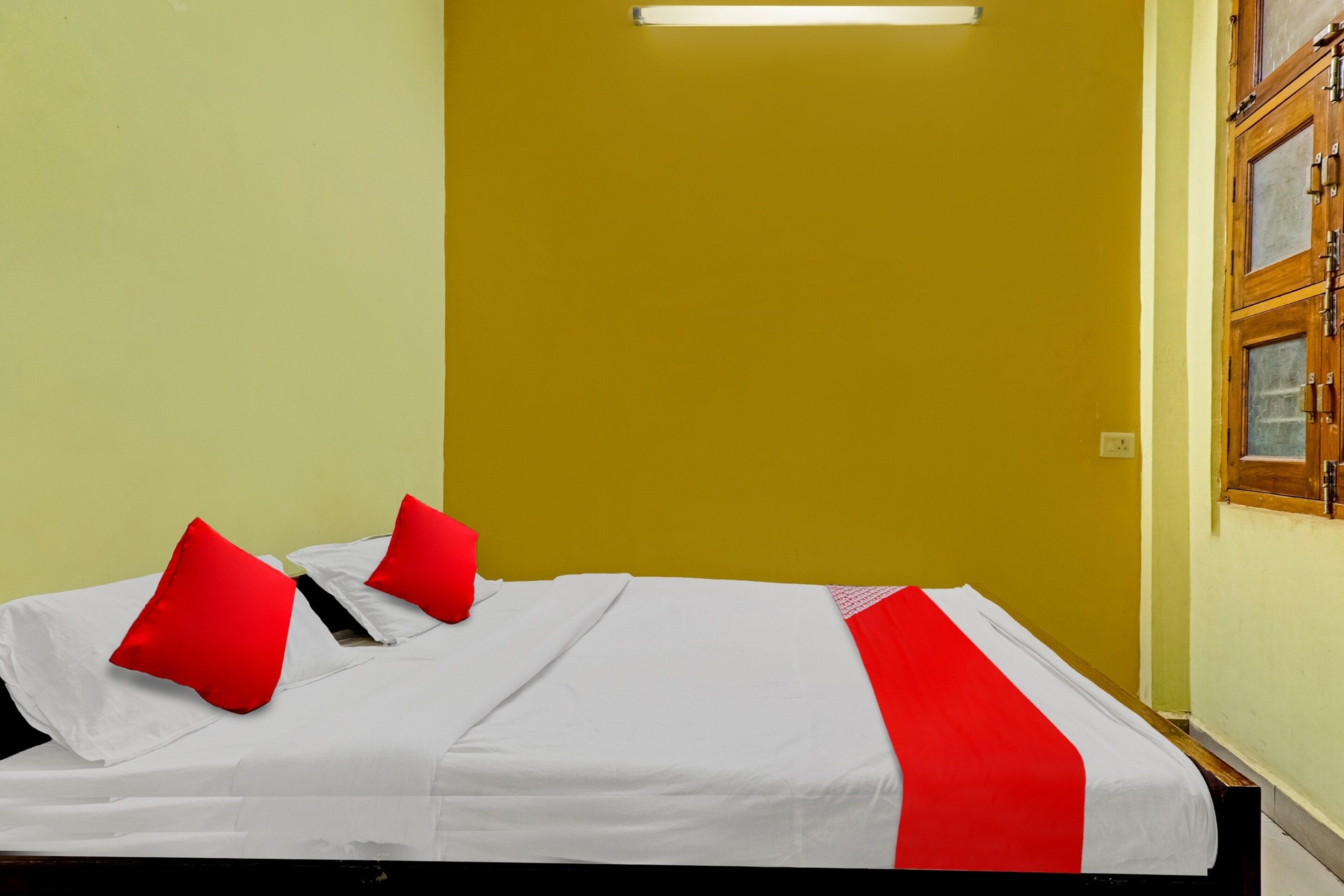 Una cama individual con ropa blanca y almohadas rojas en una habitación con paredes amarillas y una puerta de madera.