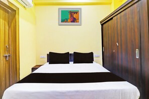 A hotel room with a bed, two black pillows, a nightstand, a wall-mounted light switch, a power outlet, a framed picture on the wall, and a wooden wardrobe. - Hotel O by OYO Heaven Inn (Gwalior)