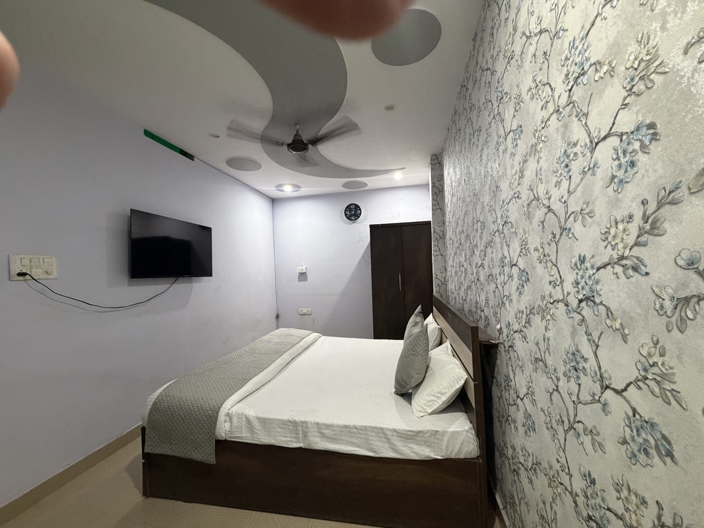 A bedroom with a bed, a flat-screen TV mounted on the wall, a ceiling fan, and a floral-patterned wallpaper on one wall.