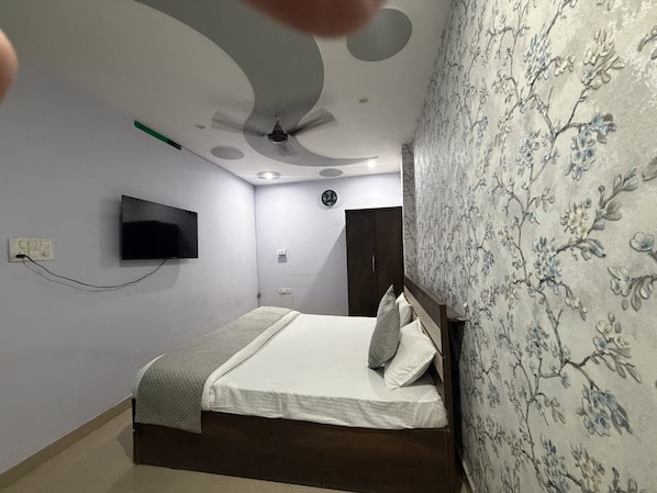 A bedroom with a bed, a flat-screen TV mounted on the wall, a ceiling fan, and a floral-patterned wallpaper on one wall. - Hotel PRAGATI INN (Kanpur)