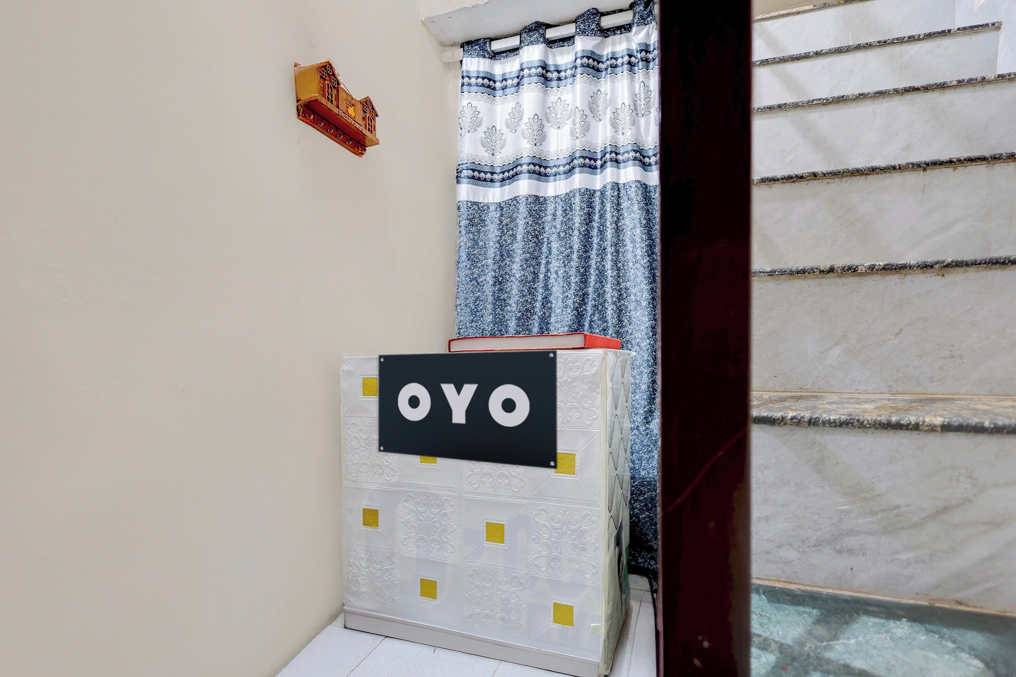 A room with a white wall, a small wooden house decoration, a patterned curtain, and a sign with 'OYO' on a white object.