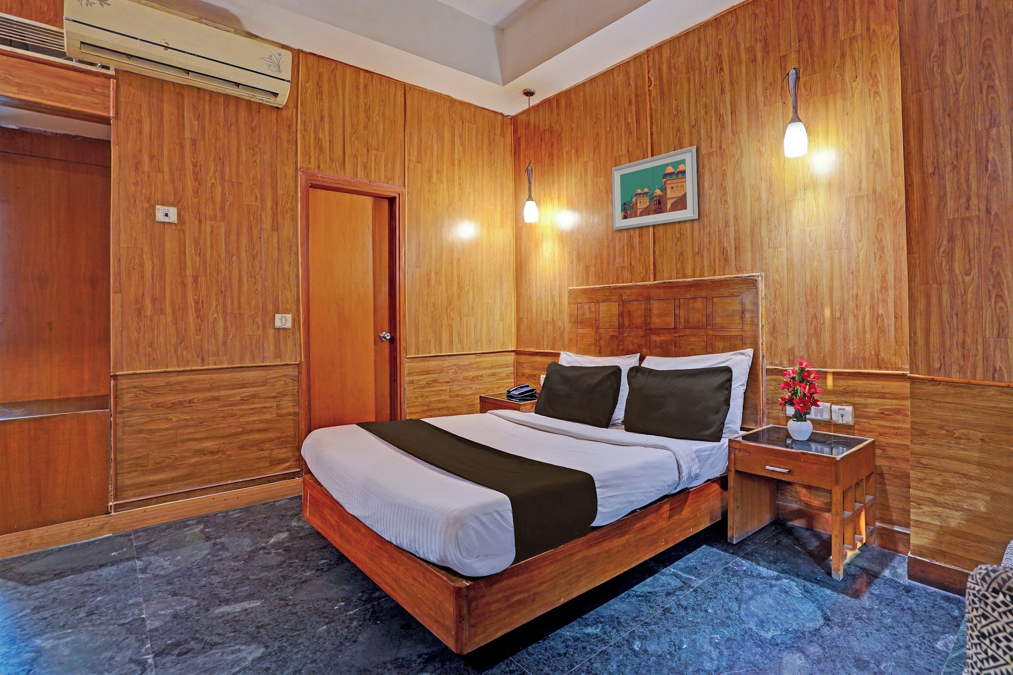 A hotel room with a wooden headboard, a bed with white bedding, a nightstand with a phone and a vase of flowers, and a picture on the wall.