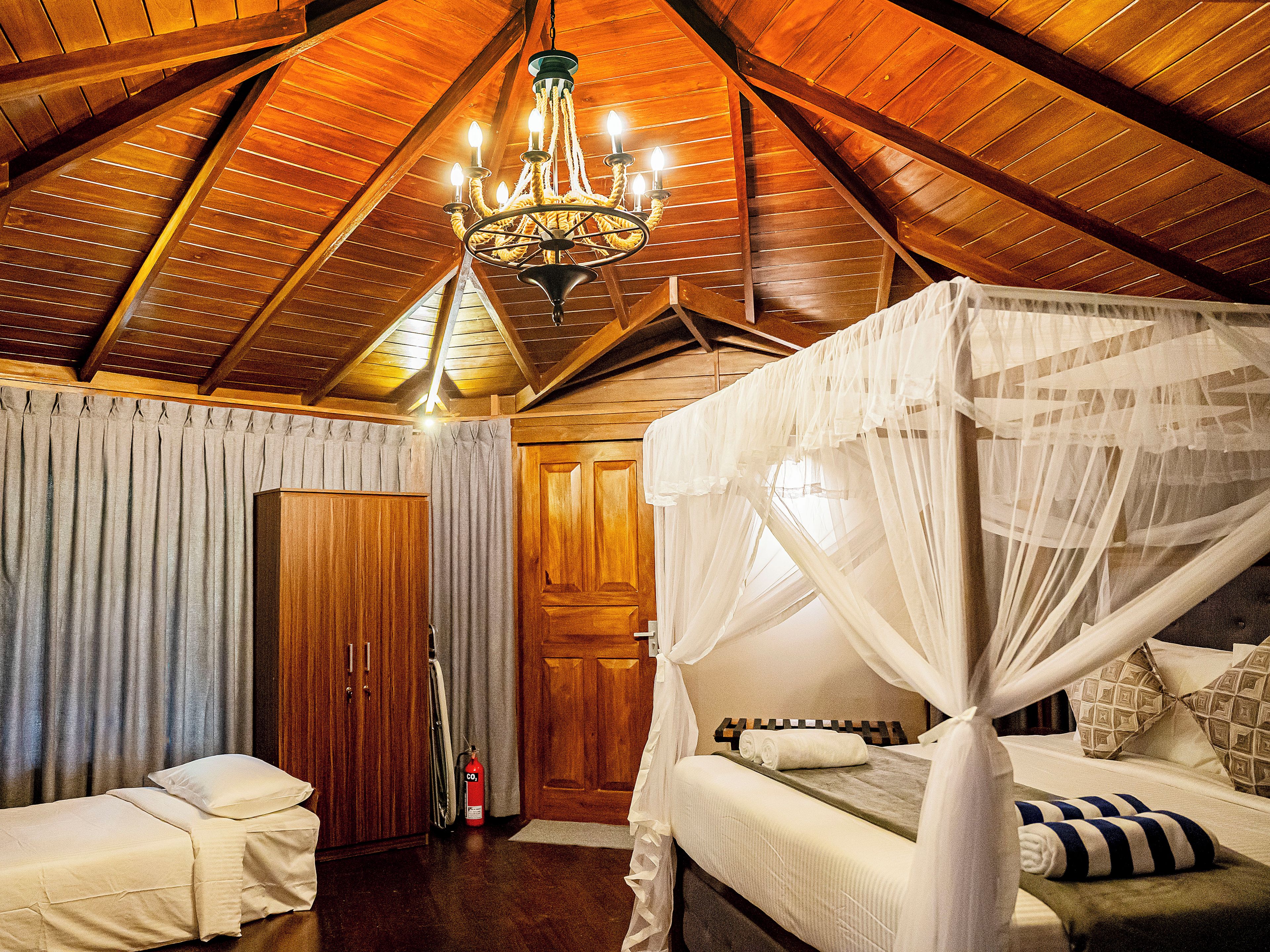 A bedroom with a wooden ceiling, a chandelier, a bed with a canopy, and a fire extinguisher.