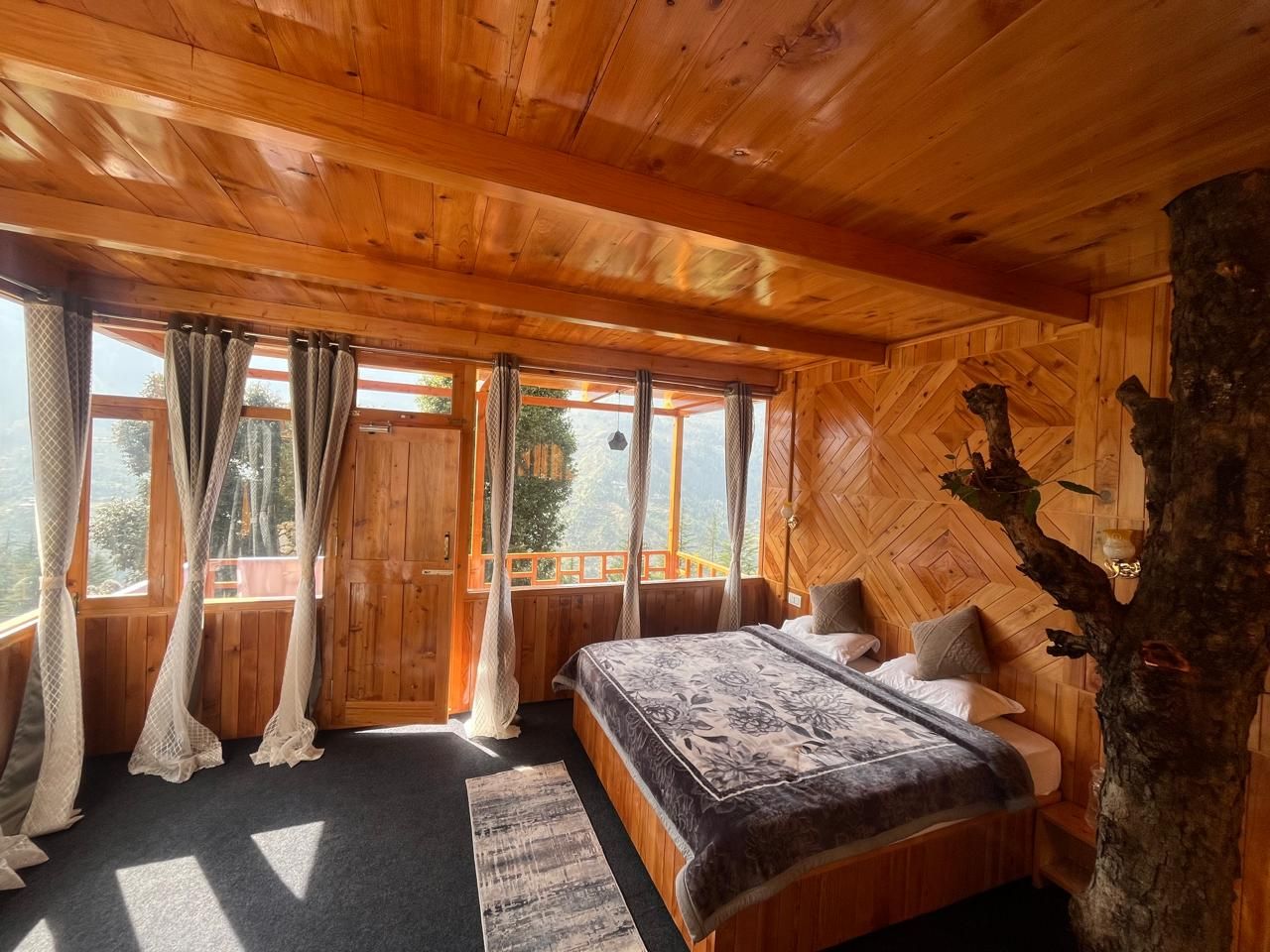 A wooden cabin bedroom with a bed, curtains, and a view through large windows.