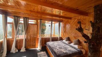 A wooden cabin bedroom with a bed, curtains, and a view through large windows.