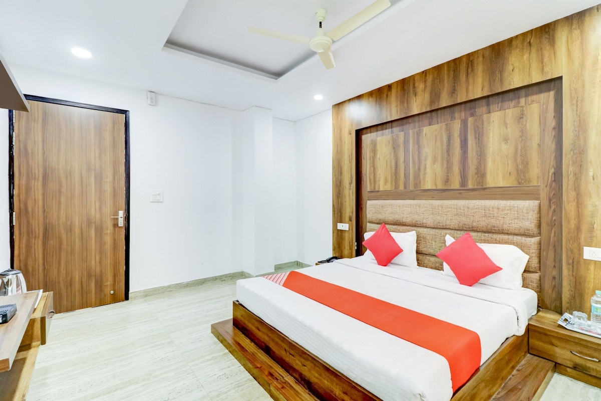 A hotel room with a wooden headboard, a bed with white bedding and red accents, a bedside table with a lamp, and a ceiling fan.