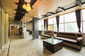Um saguão de hotel com balcão de recepção, um sofá, uma mesa de centro com flores e uma grande janela com cortinas.