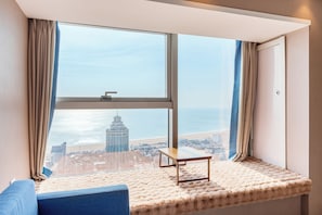 A room with a large window overlooking a beach and a cityscape, featuring a sofa and a small table. - Junyi Scenic Apt Shilaoren Intl Expo (Qingdao)