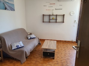 Quarto com sofá, pequena mesa de madeira, prateleira na parede com objetos decorativos e relógio.